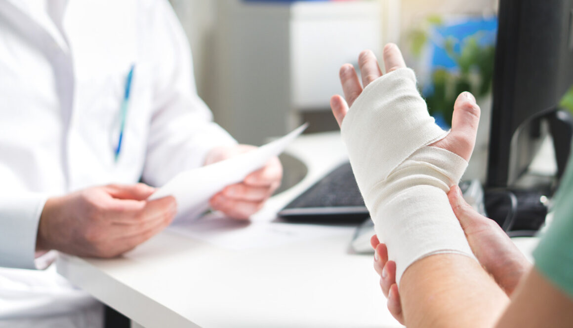 A Patient Holding a Bandaged Broken Hand on Their Doctor’s Desk Signs a Broken Bone Is Not Healing