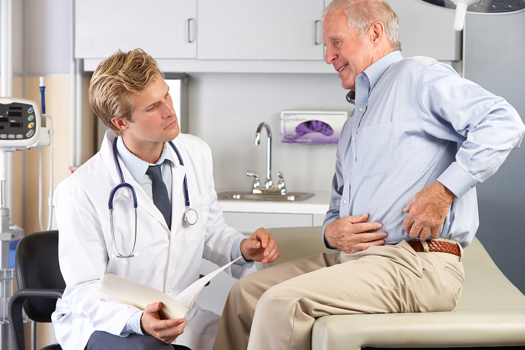 A Senior Male Patient Sitting Down Showing a Young Male Doctor Where He Is Experiencing Hip Pain How Long Does a Hip Replacement Last