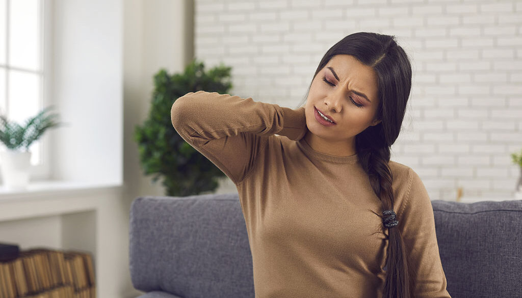 A Woman Sitting On A Couch Holding Her Neck In Pain How Long Does Whiplash Last