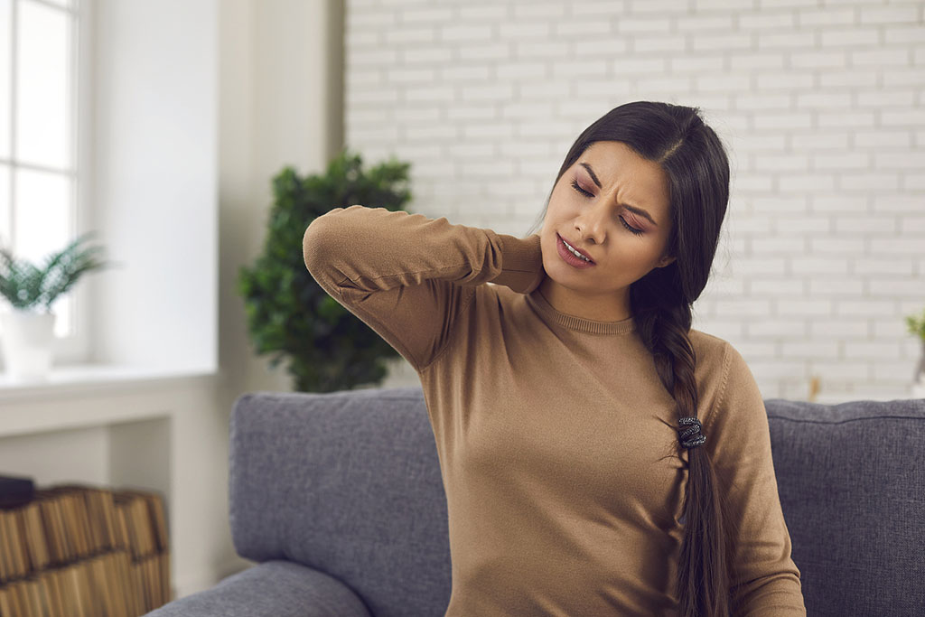 A Woman Sitting On A Couch Holding Her Neck In Pain How Long Does Whiplash Last